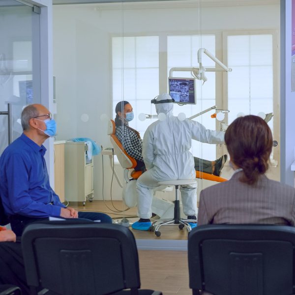 Patients with protection masks waiting doctor in dental clinic Patients with protection masks waiting doctor in dental clinic reception while stomatologist working in background wearing ppe suit. Concept of new normal dentist visit in coronavirus outbreak.